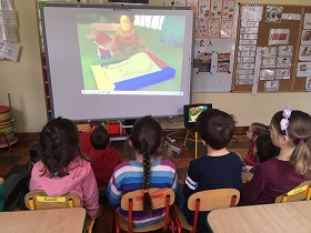 Na tablicy multimedialnej wyświetlany jest film, na którym widać chłopca przy piaskownicy. Dzieci siedzą na krzesełkach i na podłodze i wpatrują się w ekran.