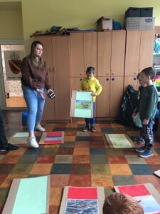 Dzieci wraz z dziewczyną stoją na środku sali i pokazują planszę ze zdjęciami. Dziewczyna trzyma w ręku czarną kostkę do gry.