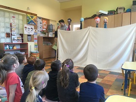 Dzieci oglądają przedstawienie kukiełek, które wystają znad białej kurtyny.