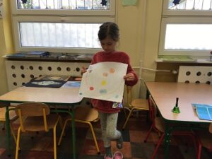 Dziewczynka stoi z rysunkiem planety w dłoniach. Za nią stoją stoliki i krzesła