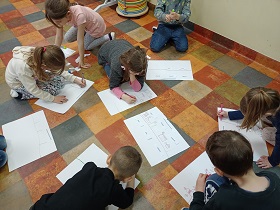 Dzieci siedzą na podłodze i mają przed sobą białe kartki papieru, na których rysują coś flamastrami.