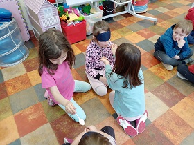 Dziewczynki i chłopcy siedzą na podłodze w parach. Dwie dziewczynki mają łyżeczki i dają je koleżankom z zasłoniętymi oczami opaską.
