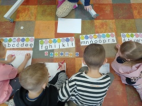 Na podłodze w sali leżą kartki z pisankami i literami. Dzieci patrzą na kartoniki i litery i piszą flamastrami na białych kartkach.