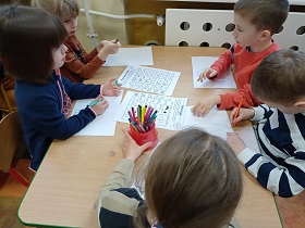 Dzieci siedzą przy stole i rysują flamastrami na białych kartkach. Na środku przed nimi leżą dwie zapisane kartki.