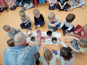 Grupa dzieci siedzi w kółku. Przed nimi leży dywanik z doniczkami. Dzieci pstrykają w nie palcami.Pomaga im pan.