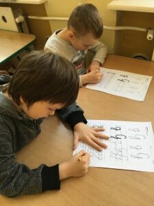 Dwoje chłopców siedzi przy stole i pisze ołówkiem literkę g w liniaturze.
