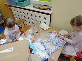 Dwie dziewczynki wycinają elementy z kolorowych gazet. Na stoliku znajdują się rolki po papierze, kartony, flamastry i talerzyki papierowe.