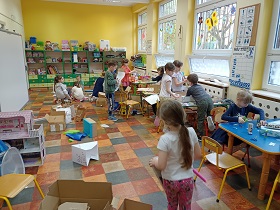 Dzieci pracują przy stolikach i na podłodze na kartonach. Wycinają i przyklejają do nich elementy.