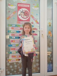 Dziewczynka stoi z dyplomem w ręku. W tle za nią są drzwi z naklejonymi kolorowymi, papierowymi kredkami i nazwą biedronki.