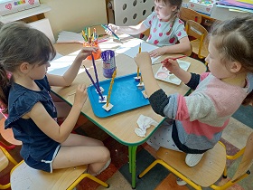 Trzy dziewczynki siedzą przy stole. Dwie z nich malują makaron farbami, jedna z nich rysuje flamastrami po białej kartce.