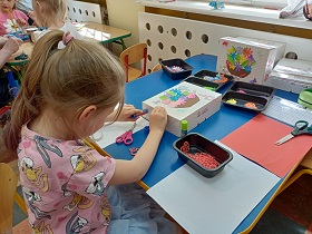 Dziewczynka przykleja klejem coś do pudełka. Na stole leżą dwa pudełka, pojemniki z kolorowymi wyciętymi elementami oraz kartki papieru.