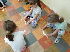Dzieci siedzą na podłodze. Przed nimi leżą białe kartoniki z literami. Dzieci je układają.