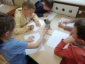 Piątka dzieci siedzi przy stole i rysuje flamastrami na białych kartkach. 