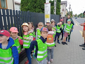 Grupka dzieci w zielonych kamizelkach, stoi na przystanku autobusowym.