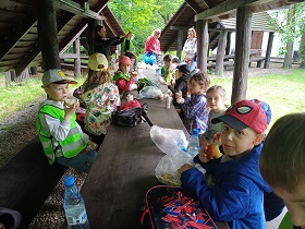 Dzieci siedzą przy długim, drewnianym stole i zjadają bułki.