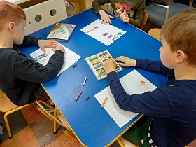 Trójka dzieci siedzi przy stolikach i rysuje pastelami na białych kartkach.