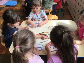 Dzieci siedzą przy stoliku i rysują kredkami na białych kartkach.
