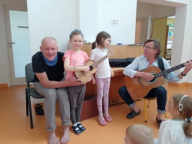 Dwie dziewczynki stoją przy dwóch panach. jeden z nich trzyma gitarę w rękach. Dziewczynka w różowej bluzce gra na strunach ukulele.