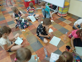 Dzieci siedzą na podłodze i wycinają nożyczkami kształty z niebieskich kartek. Przed nimi leżą puste butelki po wodzie.