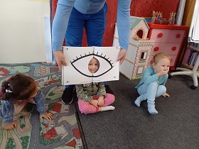 troje dzieci siedzi na dywanie, pośrodku jest dziewczynka, do twarzy Pan przyłożył książkę z narysowanym okiem.