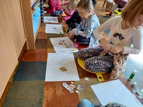 Dzieci siedzą na podłodze i przyklejają do białej kartki papieru małe elementy.
