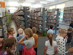 Dzieci stoją w rozsypce w sali z książkami, ustawionymi na regałach. 