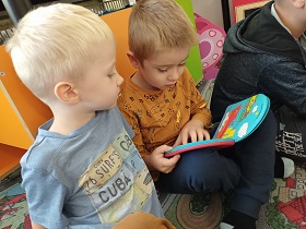Dwoje chłopców ogląda wspólnie książkę. Jeden chłopiec trzyma książkę opartą na kolanach. 