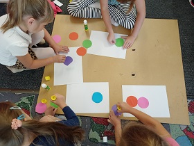 Dzieci przyklejają kolorowe kółka do białych kartek A4. Pracują na drewnianej dykcie, która leży na dywanie. 