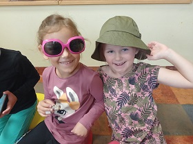 Dwie dziewczynki siedzą na podłodze. Jedna ma na nosie różowe okulary przeciwsłoneczne, a druga na głowie oliwkowy kapelusz. 