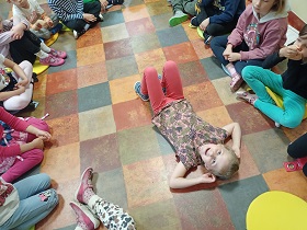 Dzieci siedzą w kółku na podłodze. Jedna dziewczynka leży w środku niego, na plecach. 