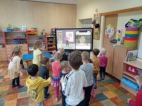 Dzieci stoją przed monitorem i wykonują pozy, które są na nim wyświetlane.