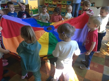 Na zdjęciu widać dzieci stojące w kółeczku, które trzymają w rękach chustę animacyjną i podrzucają ją do góry.