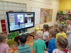 Dzieci stoją przed monitorem i spoglądają na niego. Na monitorze wyświetlany jest filmik z panią, która wykonuje ćwiczenia.