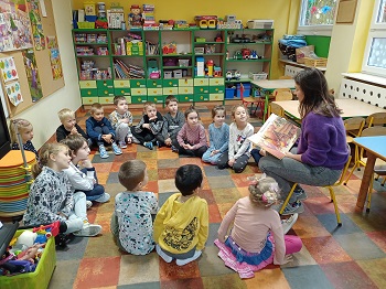 Dzieci siedzą w kółeczku w siadzie skrzyżnym, na podłodze. Razem z nimi na krześle siedzi Pani w fioletowym swetrze i trzyma książkę na kolanach. Dzieci spoglądają na panią. 