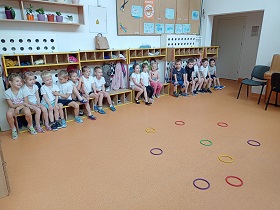 Dzieci przebrane w stroje gimnastyczne siedzą na ławkach. Przed nimi na podłodze leżą kolorowe kółka.