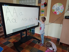 Chłopiec stoi przy monitorze i piszę literkę A. Dziewczynka za nim na niego spogląda.