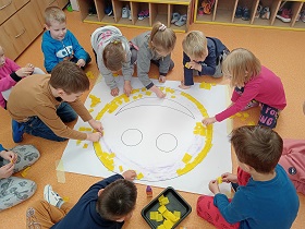 Dzieci w grupkach przyklejają żółtą krepinę do białej buźki.