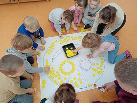 Dzieci w grupkach przyklejają żółte karteczki do białej buźki.