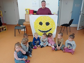Dzieci siedzą przed panią, która w rękach trzyma uśmiechniętą buzię wyklejoną żółtymi i czarnymi karteczkami. 