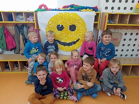 Dzieci siedzą przed uśmiechniętą buzią wyklejoną żółtymi i czarnymi kawałkami krepiny.