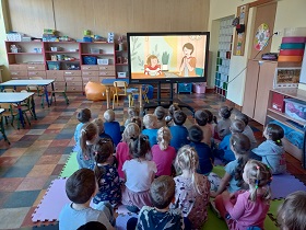 Dzieci siedzą na podłodze na matach przed monitorem. Na monitorze wyświetlana jest postać rysunkowa dziewczynki i kobiety.