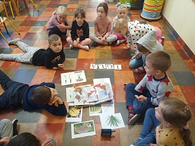 Dzieci siedzą wokół ilustracji z igłami, monografią litery I oraz słowem igła. Dwoje chłopców leży na brzuchu.