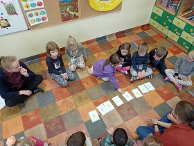 Dzieci z paniami siedzą wokół białych kartoników ułożonych w rzędzie. Dziewczynka w fioletowej bluzie wskazuje jeden z kartoników. 