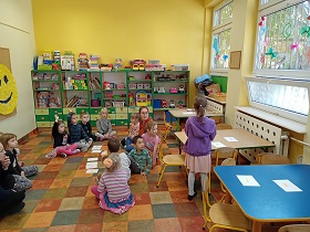 Dzieci i panie spoglądają na dziewczynkę w fioletowej bluzie, która stoi przy stoliku. 