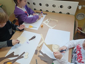 Dwóch chłopców i dziewczynka siedzą przy stoliku i wykonują pracę plastyczną. Dziewczynka i chłopiec przyklejają elementy do białej kartki A3. Drugi chłopiec wycina beżowe elementy. 