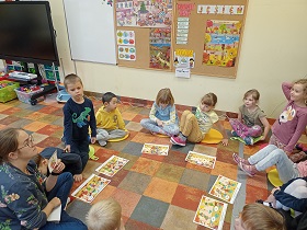 Dzieci siedzą w kółku na kolorowych poduszkach, przed nimi ułożone są ilustracje. Jedna dziewczynka się zgłasza. Pani wskazuje jej kartonik z ilustracją. 