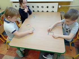 Dwóch chłopców i dziewczynka układają na stoliku z kolorowych patyczków dwa kwadraty.