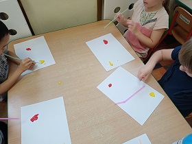 Dzieci przy stolikach siedzą i trzymają w rękach słomki. Na stoliku przed nimi leżą kartki z kleksami farby. Dzieci w nie dmuchają. 