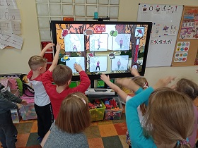 Dzieci stoją przed monitorem i wykonują ćwiczenia, które wykonuje na monitorze pani.