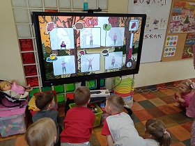 Dzieci stoją przed monitorem i wykonują ćwiczenia, które wykonuje na monitorze pani.